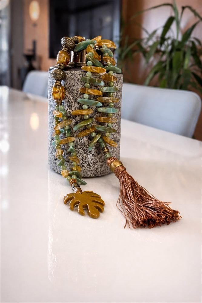 Colar de folhas de vidro âmbar e verdes com pingente em folha e borla acobreada, disposto sobre vaso de pedra granulada.