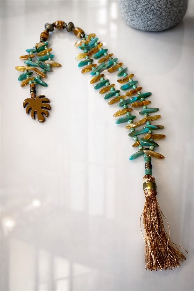 Colar de folhas de vidro em tons de água e âmbar com pingente em forma de folha e borla dourada, sobre superfície branca.