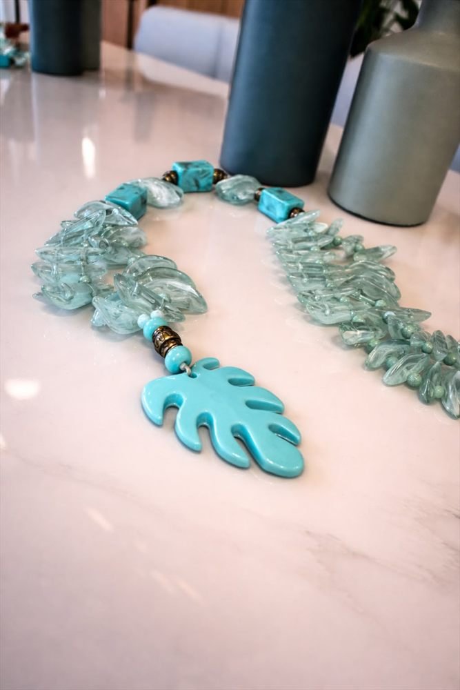 Colar de folhas de vidro em tons de água, com peças turquesa e pingente em forma de folha, sobre mesa de mármore.